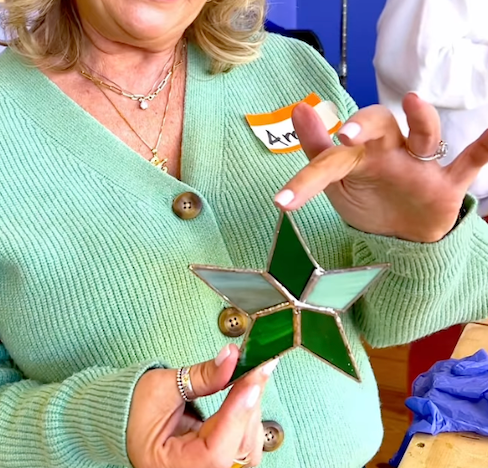 Photo of a student holding up a stained glass piece she created during a workshop. The piece is a green and white star. 