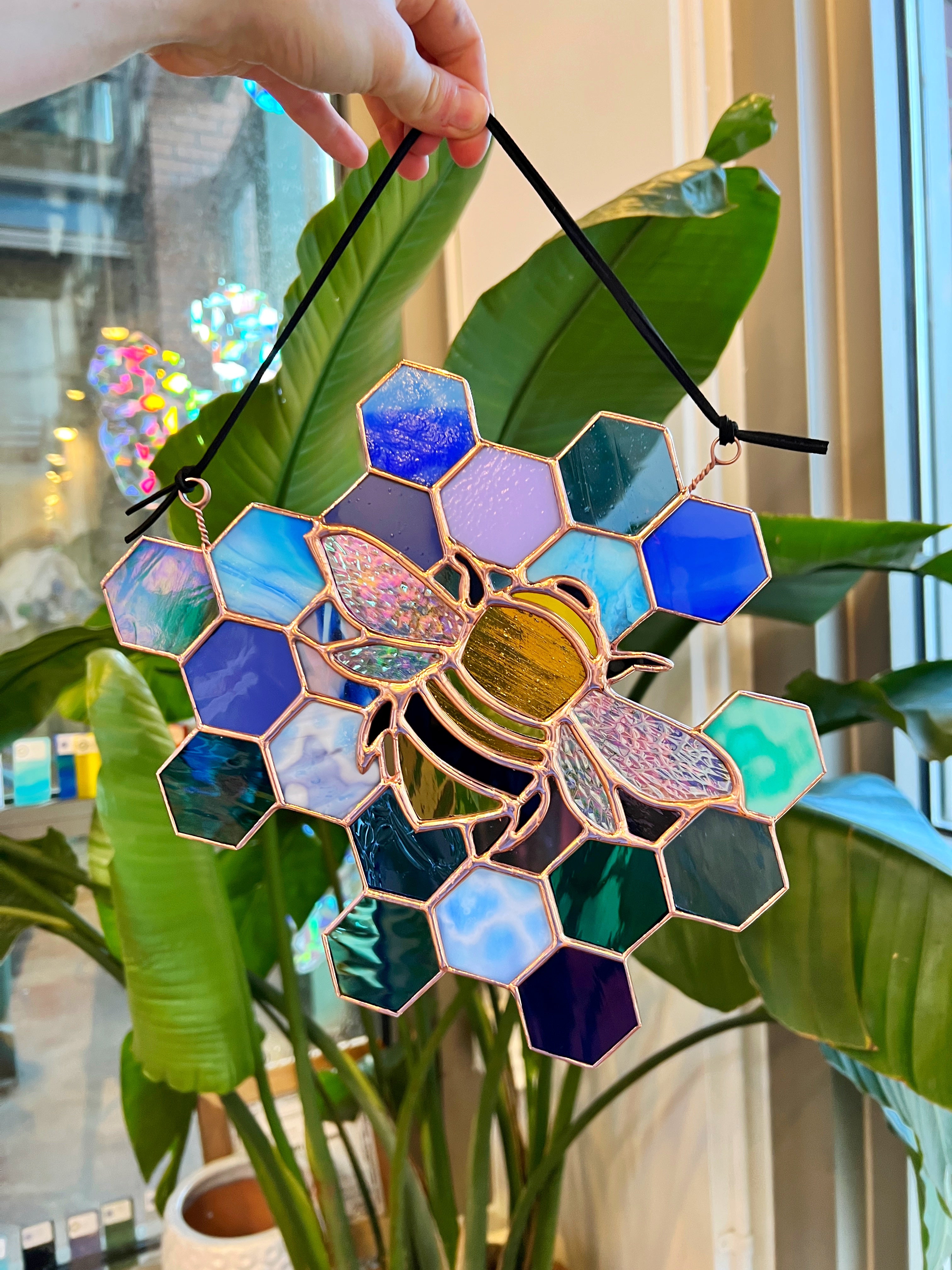 A stained glass bee suncatcher  in gold lead lines held up against green plant background.