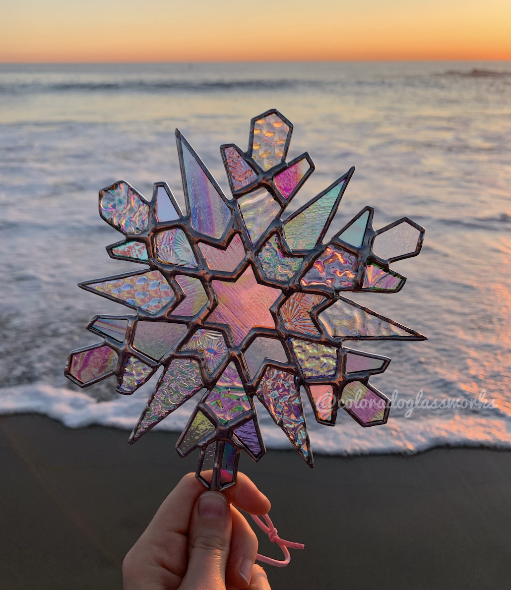 A rainbow colored stained glass snowflake against a sunsetting oceanbackground
