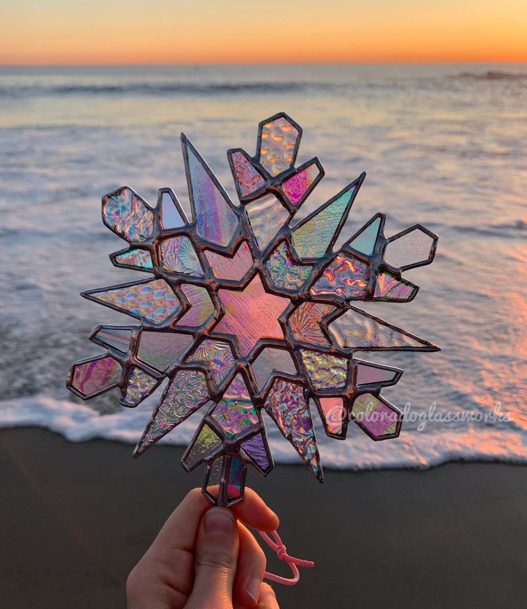 A rainbow colored stained glass snowflake against a sunsetting oceanbackground